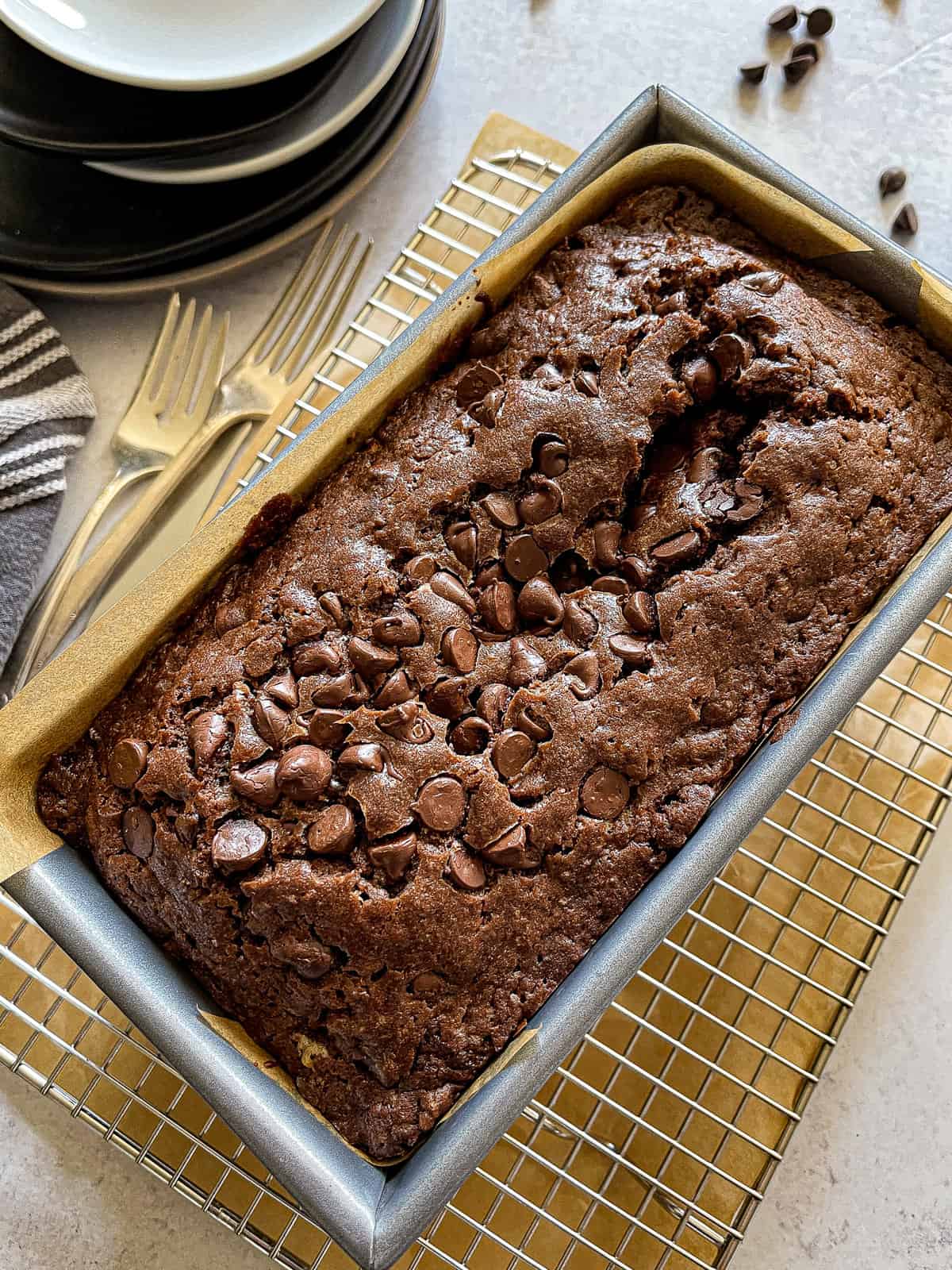 loaf of chocolate chocolate chip bread on a cooling rack, flanked by serving forks, plates, and a dark gray cloth napkin
