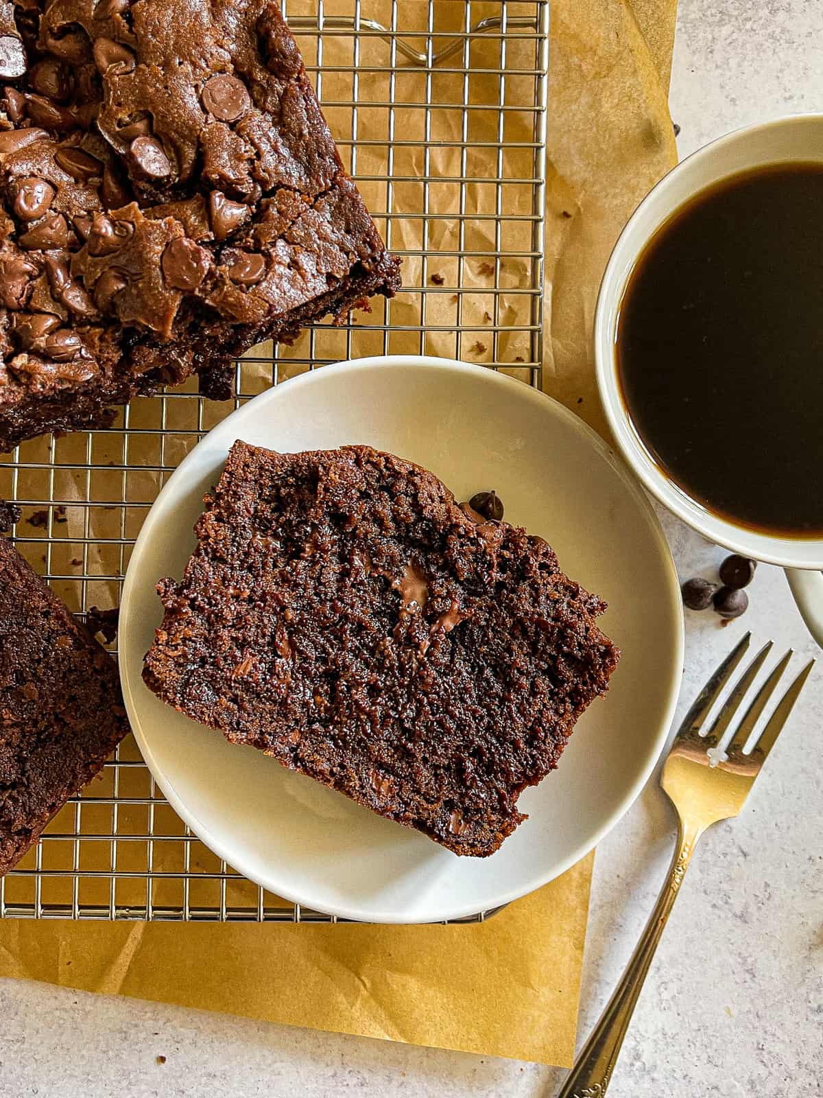 slice of chocolate chocolate chip bread on a cooling rack, flanked by serving forks, plates, and a cup of black coffee
